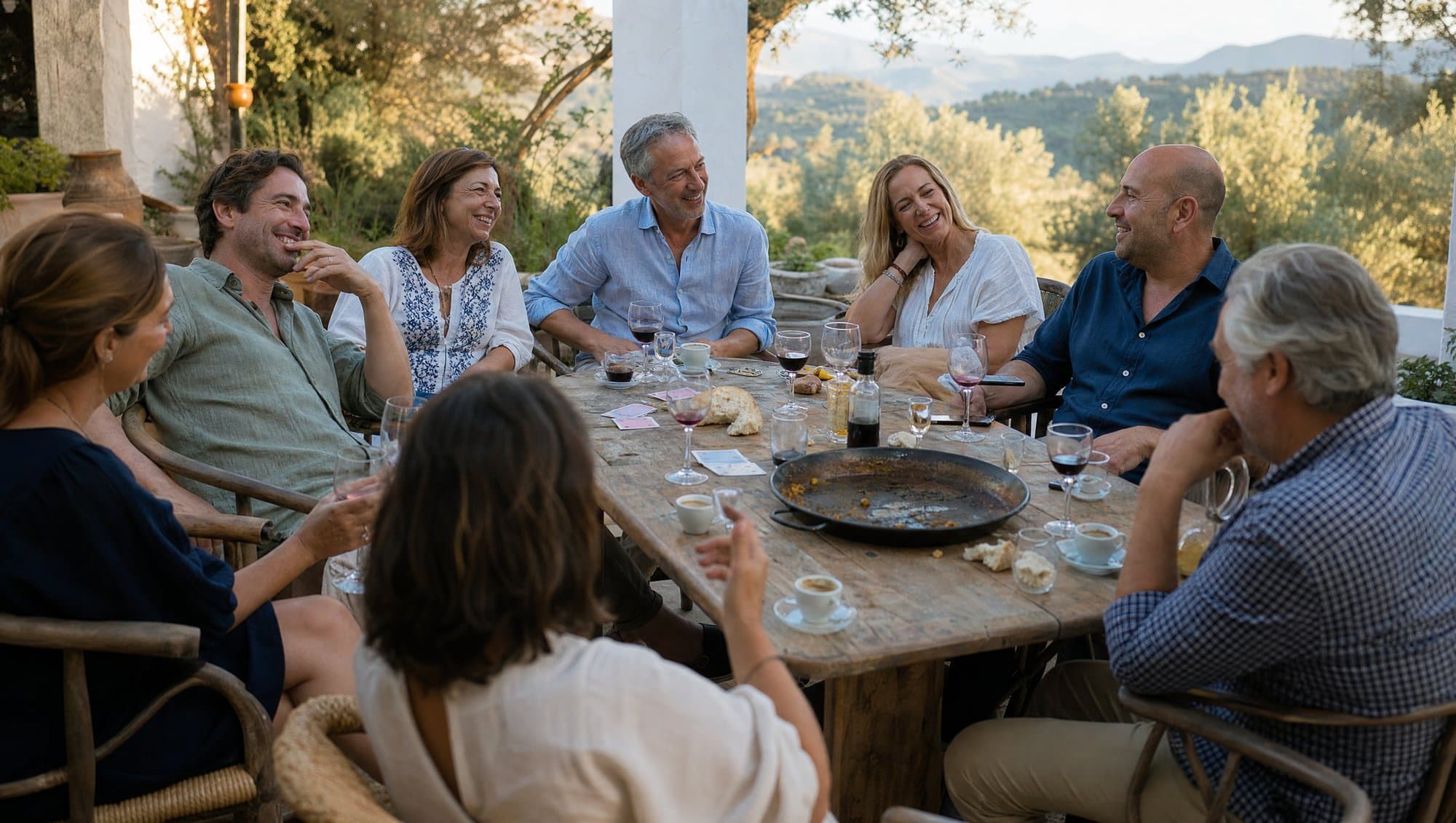 Friends lingering at a long table on a villa terrace after a Spanish lunch, with an empty paella pan, coffee cups and small glasses of sweet wine